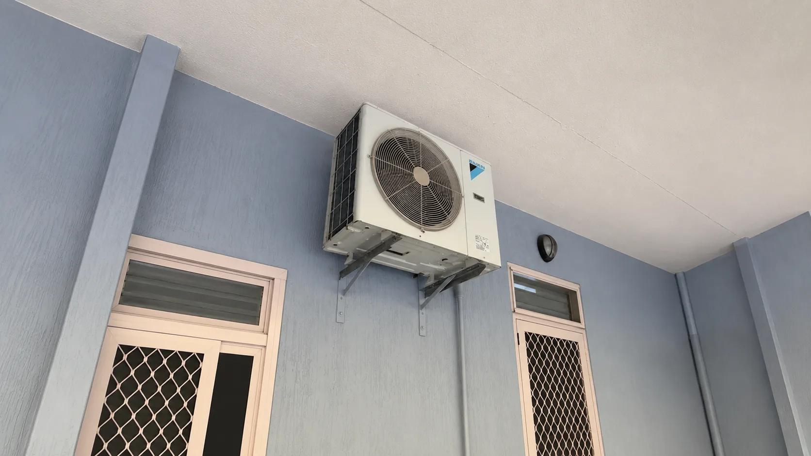 Daikin split system outdoor unit wall-mounted on steel brackets under an eave at a Coffs Harbour property, with the fan grille and Daikin badge visible and security-screened windows either side